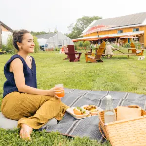 A woman sits on a picnic blanket at a brewery with farm fresh food. 