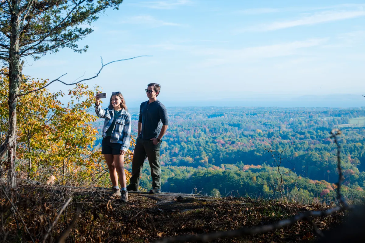 A woman and a man pose for a selfie on a mountain summit, with views of a valley in the background.