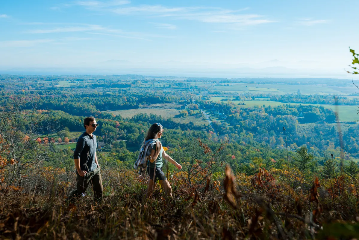 A man and woman walk a trail on a mountain in fall overlooking farm land.