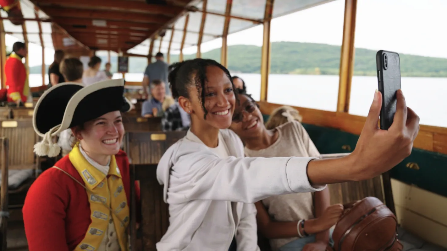 Two women pose for a selfie on a historic boat with a costumed reenactor.