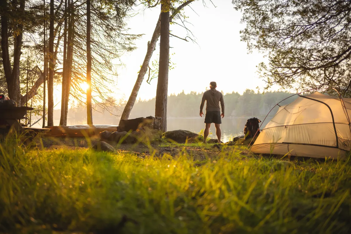 An adult stands next to a tent on a grassy campsite at sunrise.