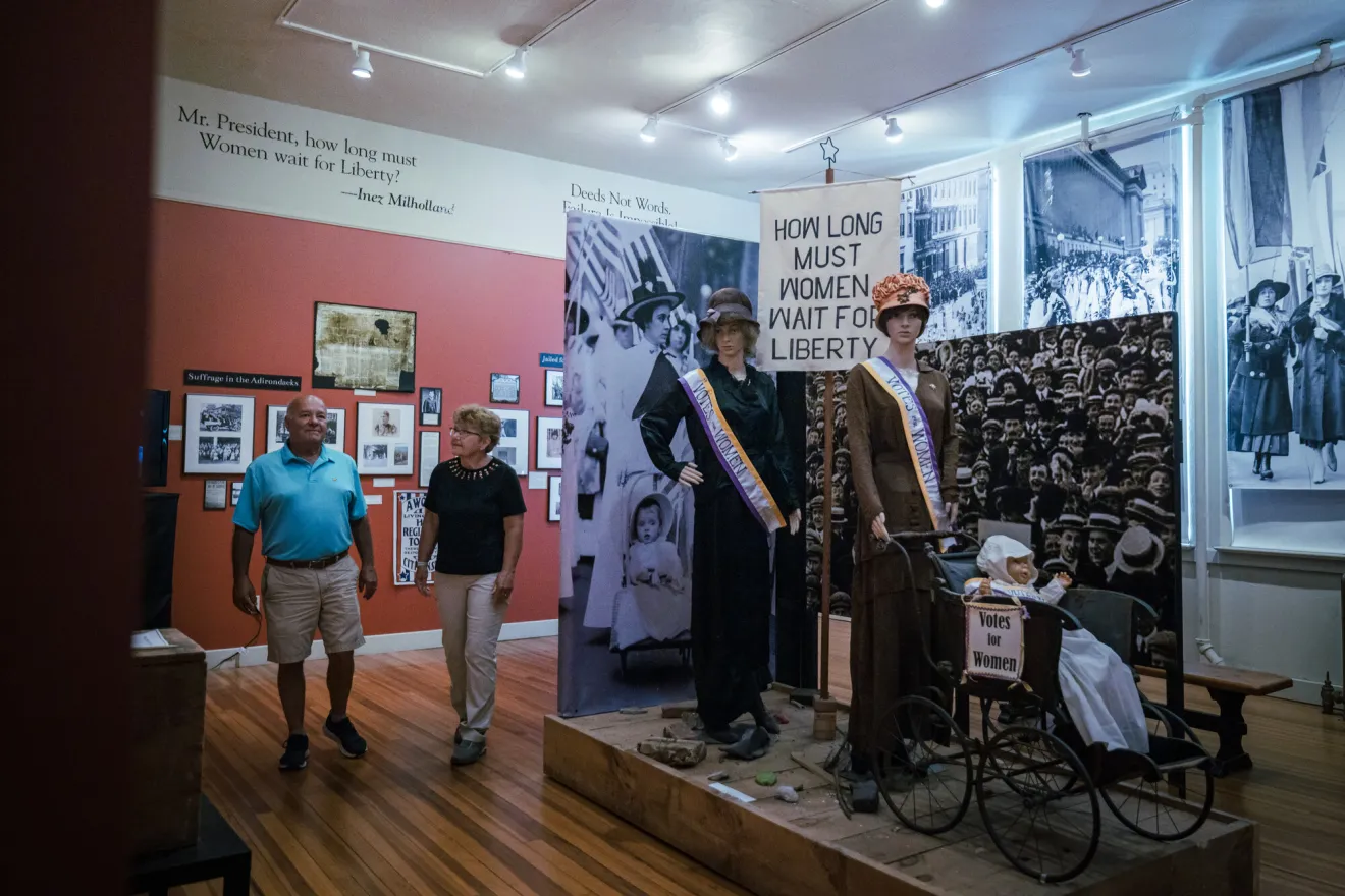 A man and a woman walk through a history museum. A display on women's suffrage is on the right.