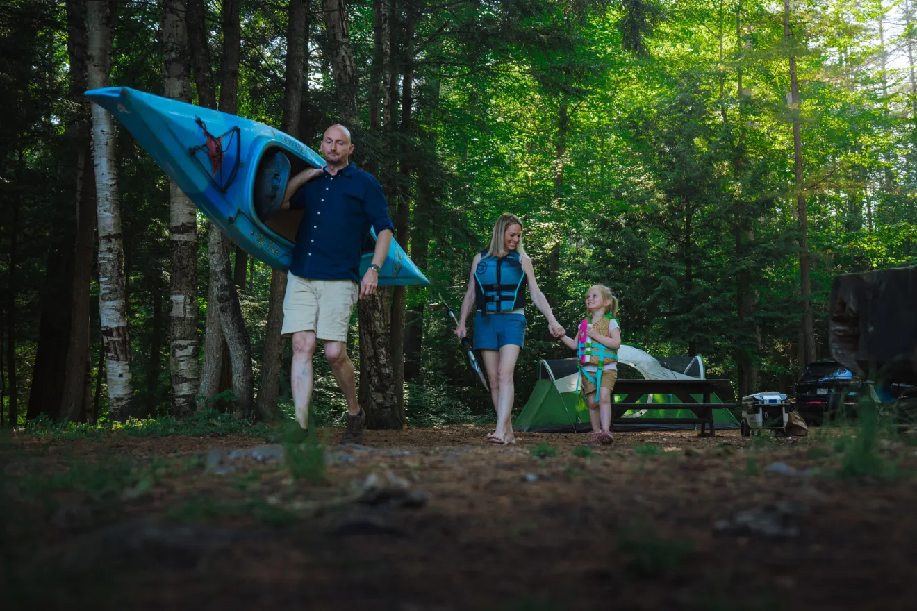 A man walks a kayak to a lake while his wife and young daughter walk behind him.