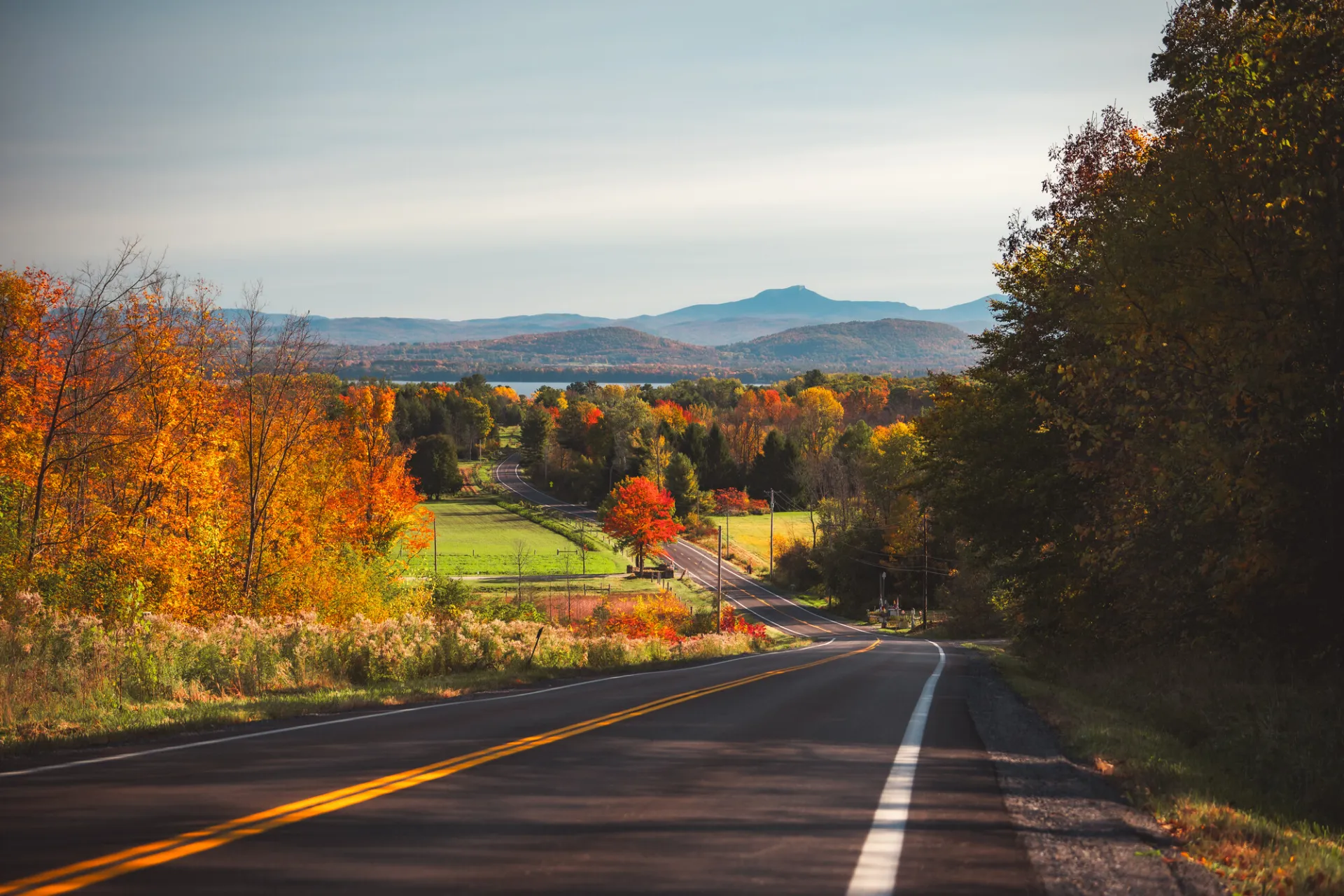 A two-lane country road runs downhill amid fall foliage-covered trees, with farmland and low mountains in the distance.
