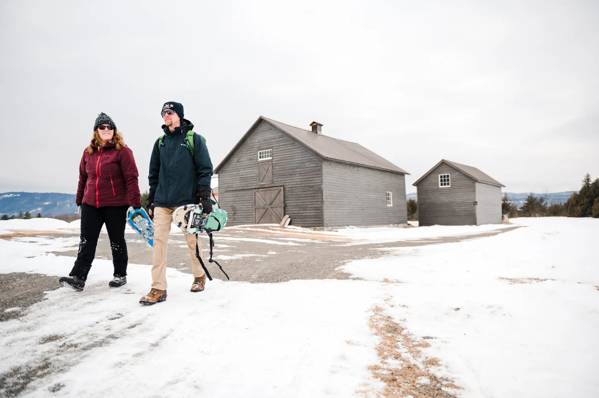 A man and woman with snowshoes in their hands walk past a gray barn in winter.