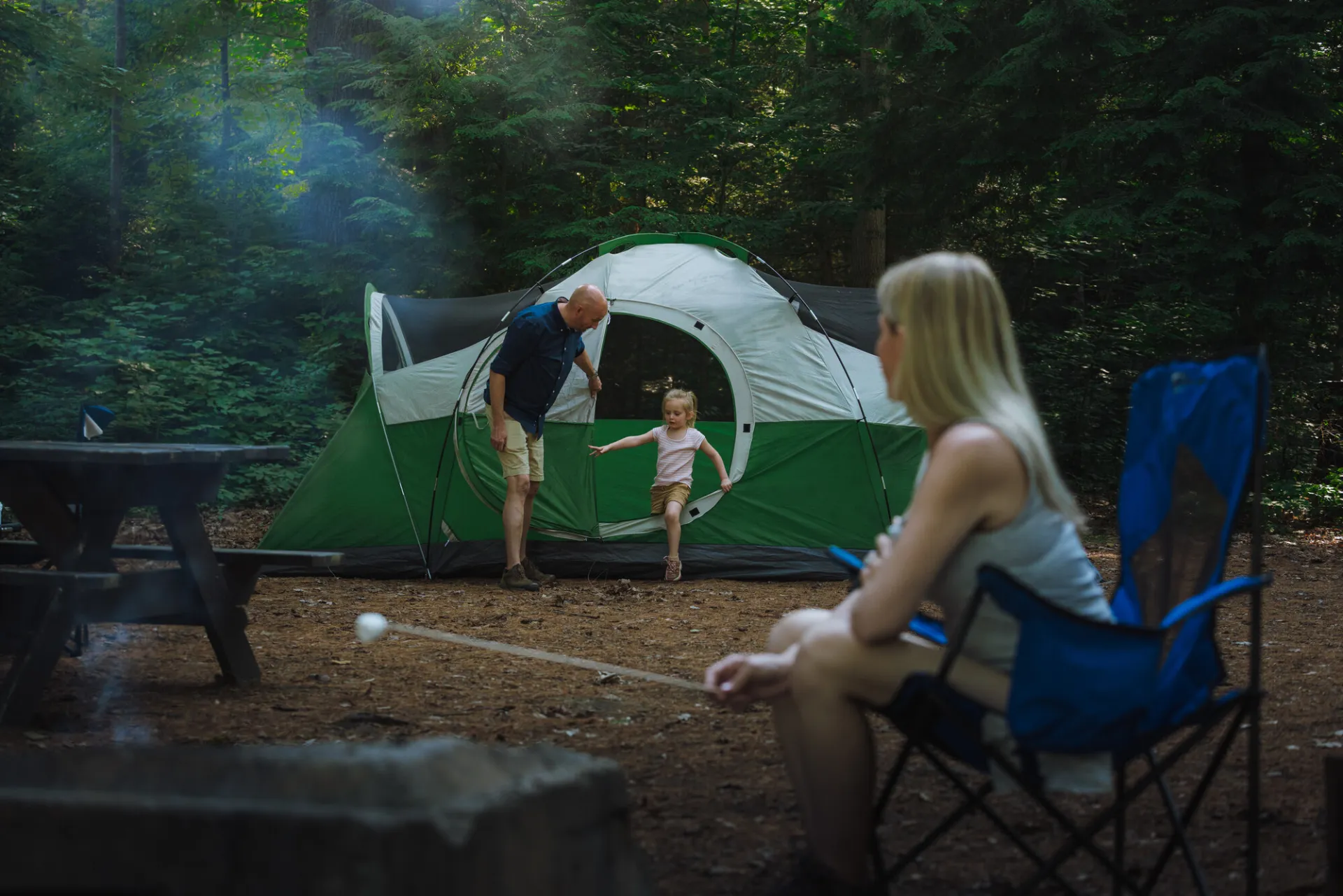 A man helps his young daughter climb out of a tent while her mom looks on while roasting marshmallows.