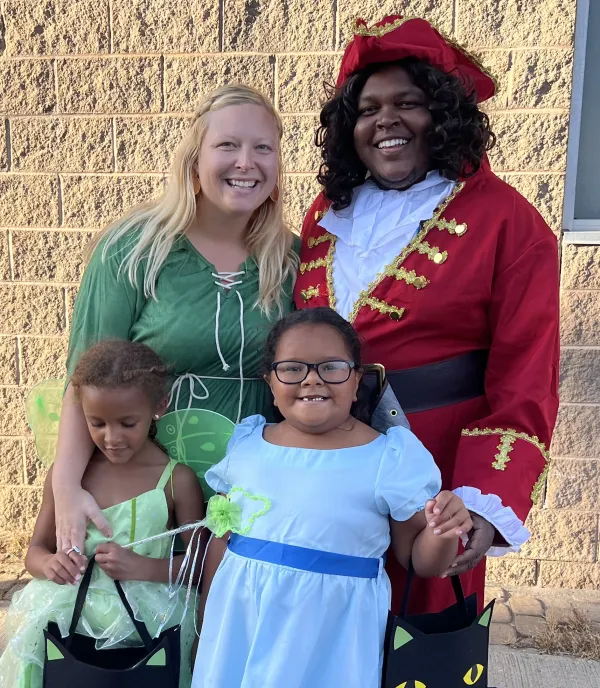 Four people in Halloween costumes. Two adults are flanking two children. The adult on the left is dressed as Peter Pan. The adult on the right is dressed as Captain Hook. One child is dressed as Tinkerbell and the other as Wendy Darling.