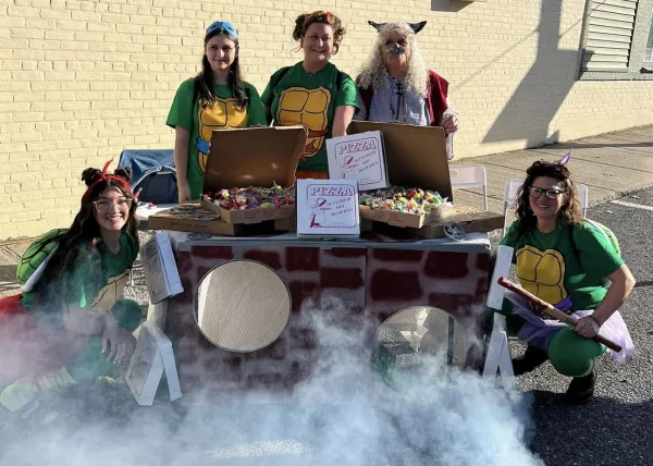 Four women dressed as Teenage Mutant Ninja Turtles and one woman dressed as a witch stand behind a table decorated as a pizza oven, which is emitting smoke.  The pizza boxes on the table are filled with candy.