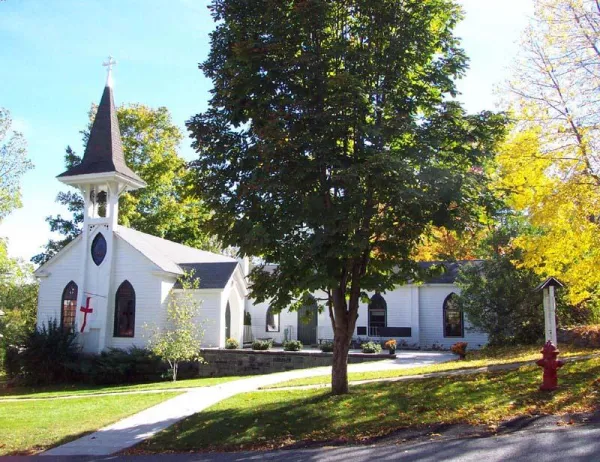A white church with grey steeple and a large pine tree out front.