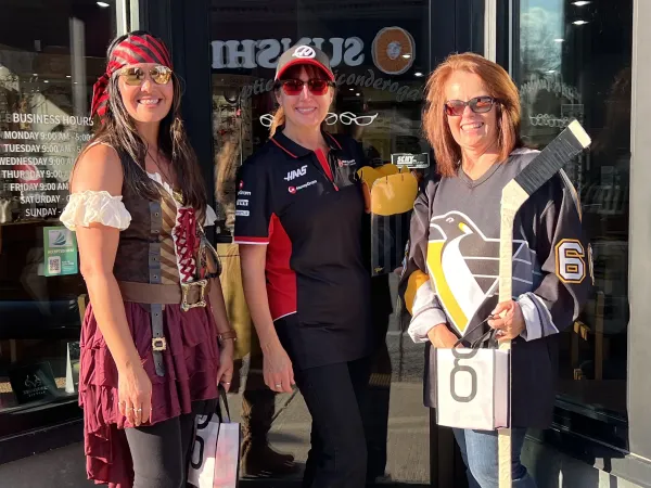Three women in costume stand outside a store. One is dressed as a pirate, one is wearing a racing team shirt, and one is wearing a Pittsburgh Penguins hockey jersey and holding a hockey stick.