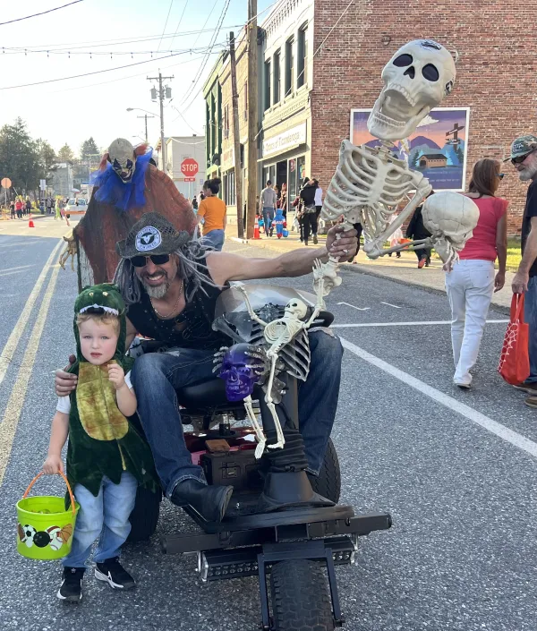A man in a cowboy hat and sunglasses sits on a motorized cart decorated with skeletons and skulls at a Halloween event, alongside a toddler in a dinosaur costume.