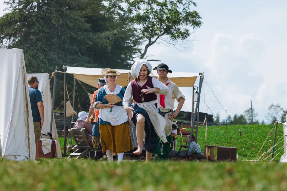 A child in colonial clothing laughs and runs toward the camera while her parents walk in the background in a field.