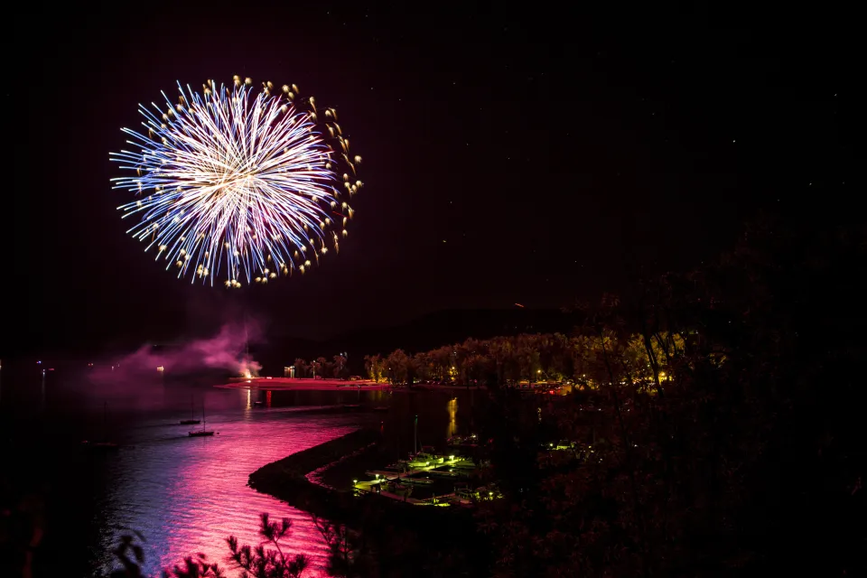 An enormous firework explodes over a small lakeshore town, the firework reflecting in the water.
