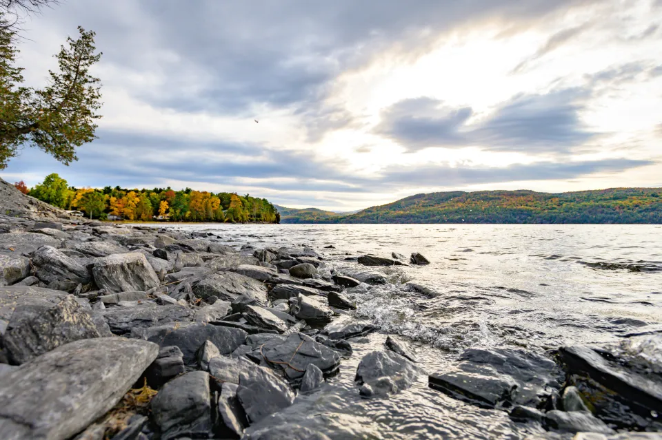 A view of Lake Champlain, with a rocky shoreline in the foreground and brilliantly colored fall foliage on the distant shore